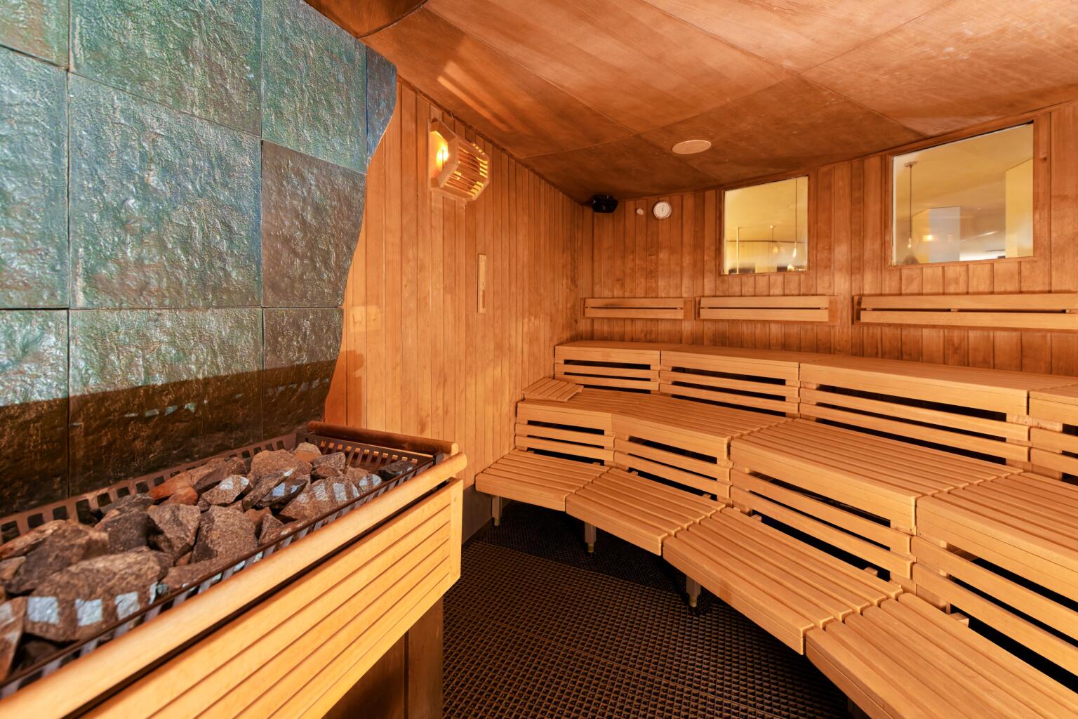 Alt-Text (detailliert): Innenraum der Meditations-Sauna mit hellen Holzwänden und gestaffelten Sitzbänken auf mehreren Ebenen. Links im Vordergrund steht ein Saunaofen mit dunklen Steinen, dahinter eine grünlich schimmernde Natursteinwand. Rechts an der Wand sind zwei Fenster zum Vorraum sichtbar, darüber warme Beleuchtung. Der Boden ist dunkel gefliest.