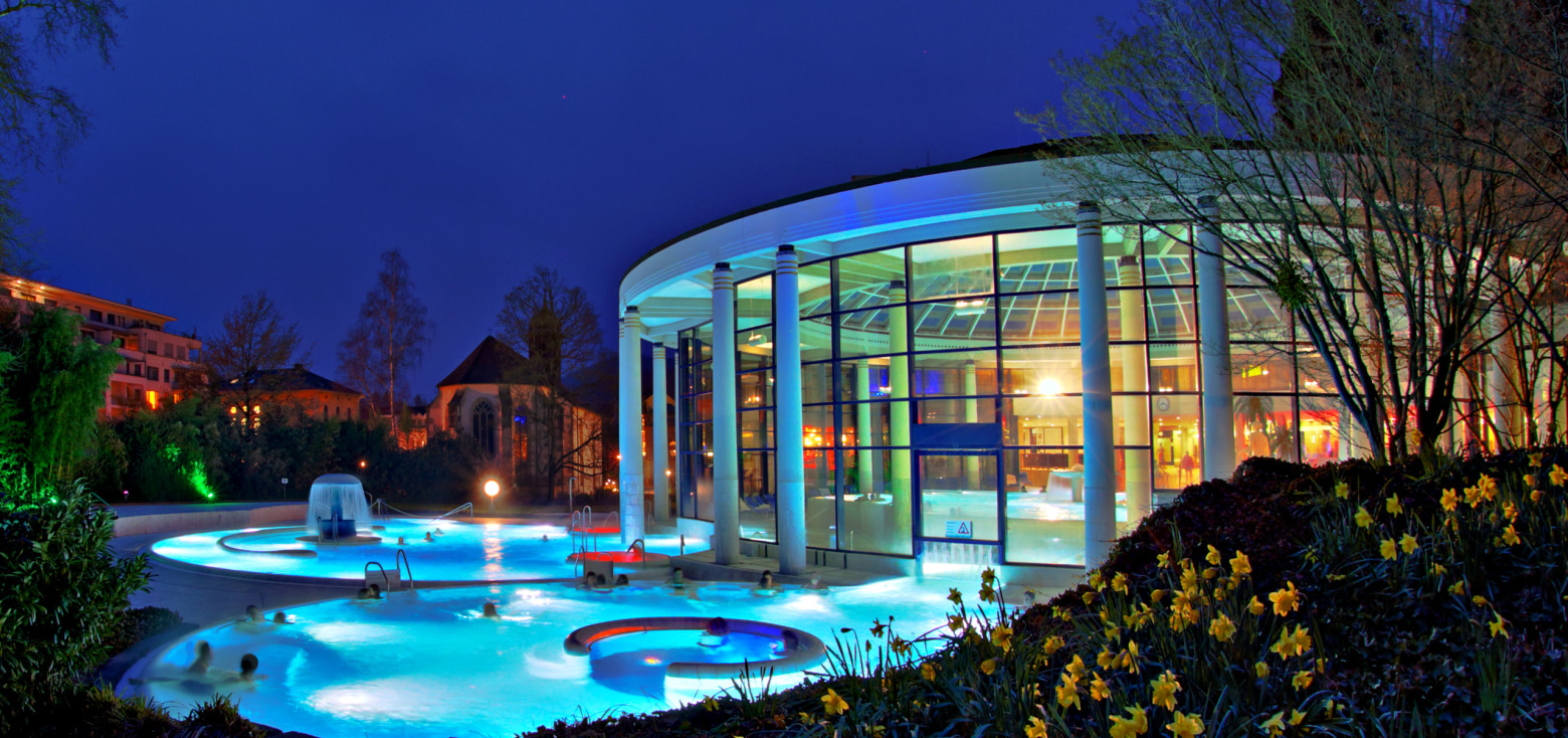 Abendaufnahme der Caracalla Therme in Baden-Baden mit beleuchtetem Außenbecken und moderner Glasfassade, umgeben von blühenden Narzissen im Vordergrund.