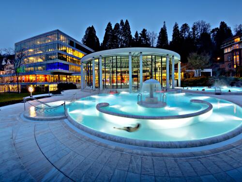 Beleuchtetes Außenbecken der Caracalla Therme Baden-Baden in der Abenddämmerung mit warmem Thermalwasser, Springbrunnen und moderner Glasarchitektur im Hintergrund.