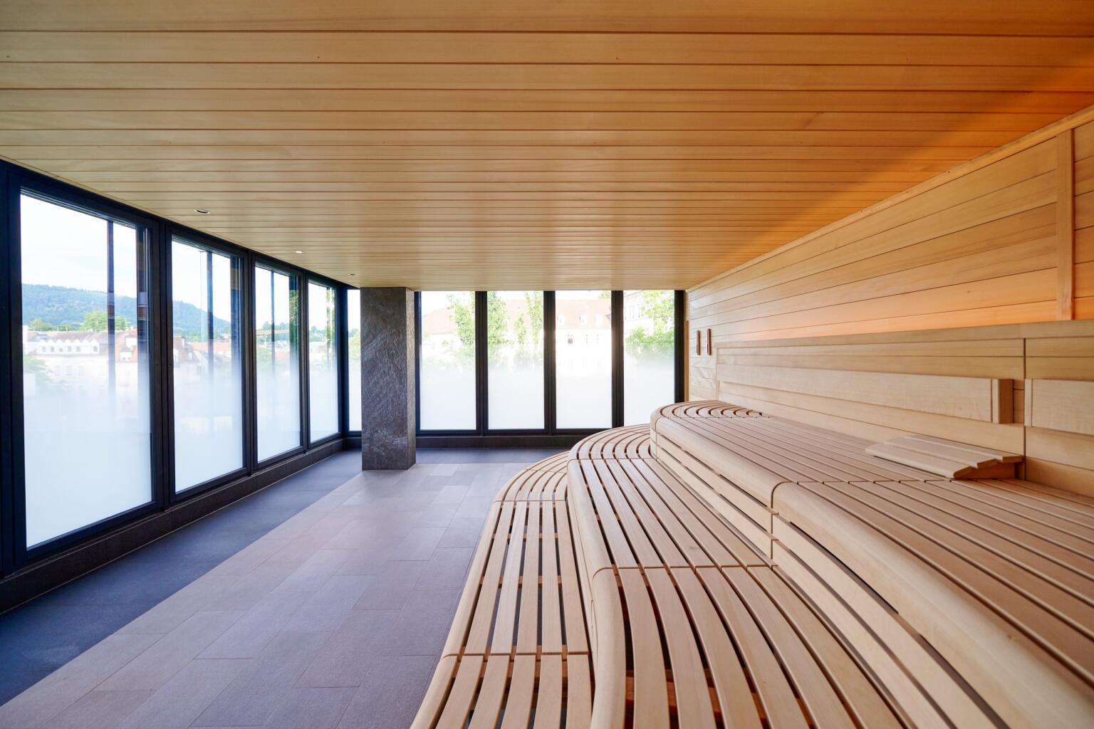 Helle Panorama-Sauna GreatSpa mit großen Glasfronten und weitem Blick über Baden-Baden. Breite, moderne Holzbänke bieten viel Platz für entspanntes Saunieren bei angenehmen 65 Grad. Ein innovativer Kombi-Saunaofen sorgt für ein sanftes Wärmeklima, das kreislaufschonend und tiefenwirksam wirkt – ideal für nachhaltige Entspannung und Wohlbefinden.