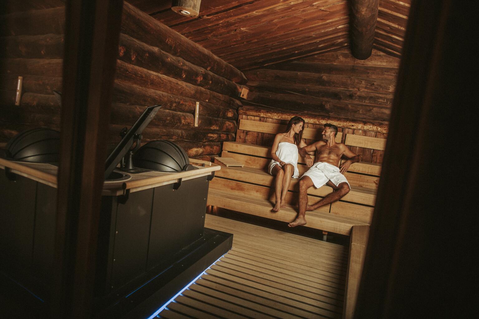 Innenraum einer rustikalen Waldsauna mit Wänden und Decke aus dunklen Holzstämmen. Links im Vordergrund steht ein moderner schwarzer Saunaofen. Auf der rechten Seite sitzen eine Frau und ein Mann in weiße Handtücher gehüllt entspannt auf gestaffelten Holzbänken und blicken sich an.