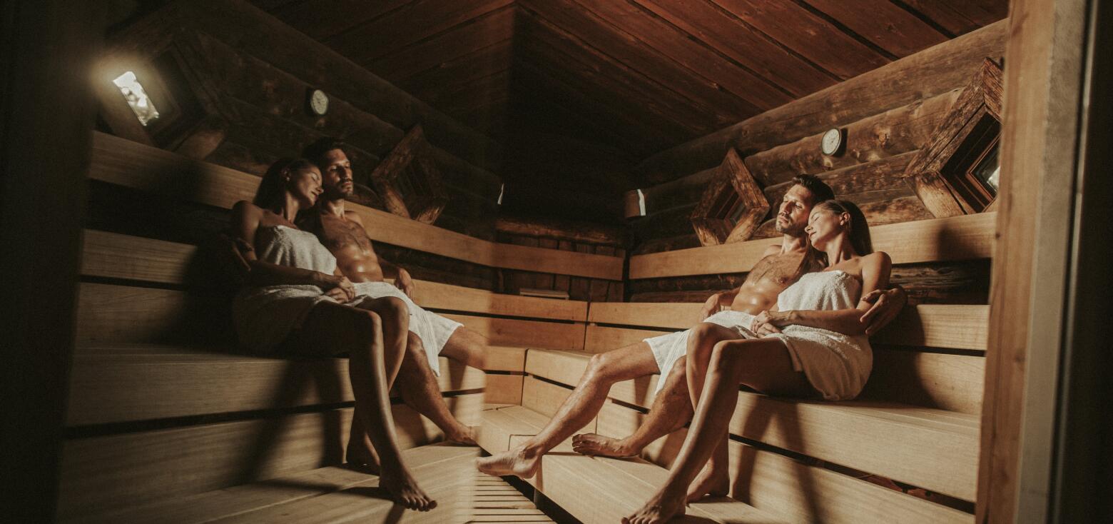 Innenansicht einer Waldsauna mit rustikaler Holzverkleidung. Zwei Paare sitzen entspannt in Handtücher gehüllt auf breiten Holzbänken und genießen die Wärme. Gedämpftes Licht und natürliche Materialien schaffen eine gemütliche, naturnahe Atmosphäre für erholsame Saunagänge.