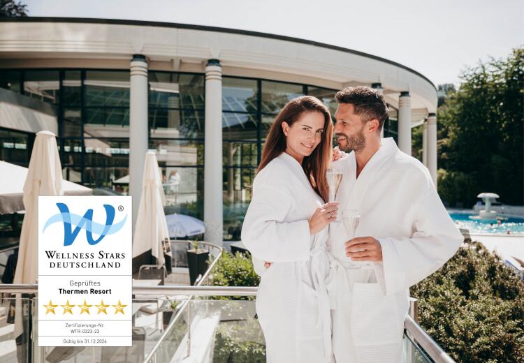 Ein Paar in weißen Bademänteln steht eng beieinander und lächelt auf der Terrasse der Caracalla Therme. Im Hintergrund die Glasfassade des Thermenresorts, eingeblendet das 5-Sterne-Siegel „Wellness Stars Deutschland