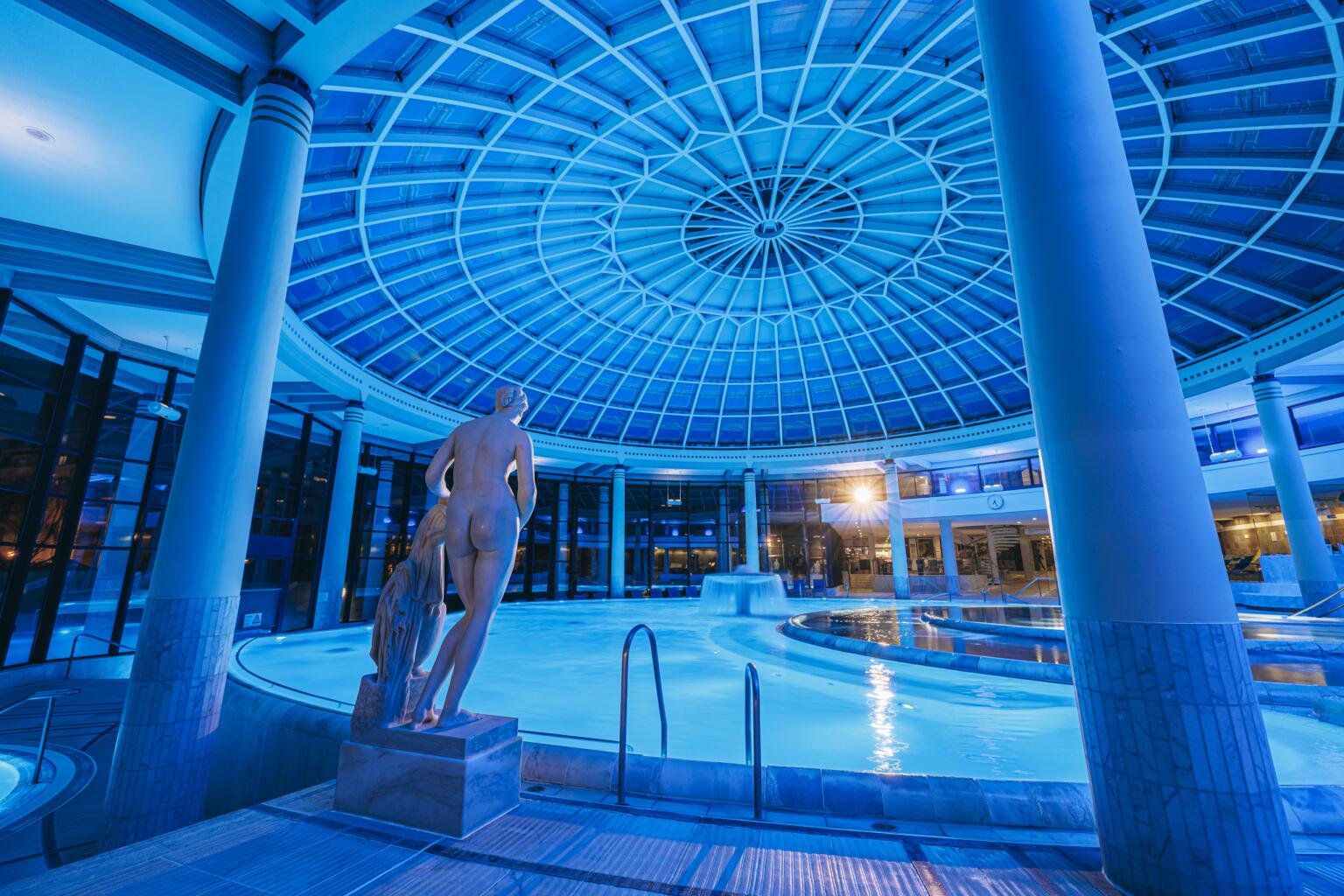Blick in die Caracalla Therme Baden-Baden: Statue im Vordergrund, dahinter ein rundes Thermalbecken unter einer großen Glaskuppel, in blauem Licht beleuchtet.